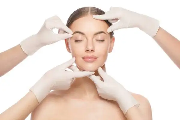 Close-up portrait of young woman standing with naked shoulders and neck, face is touched by beauticians in gloves, preparing her for plastic surgery procedures, isolated on white background